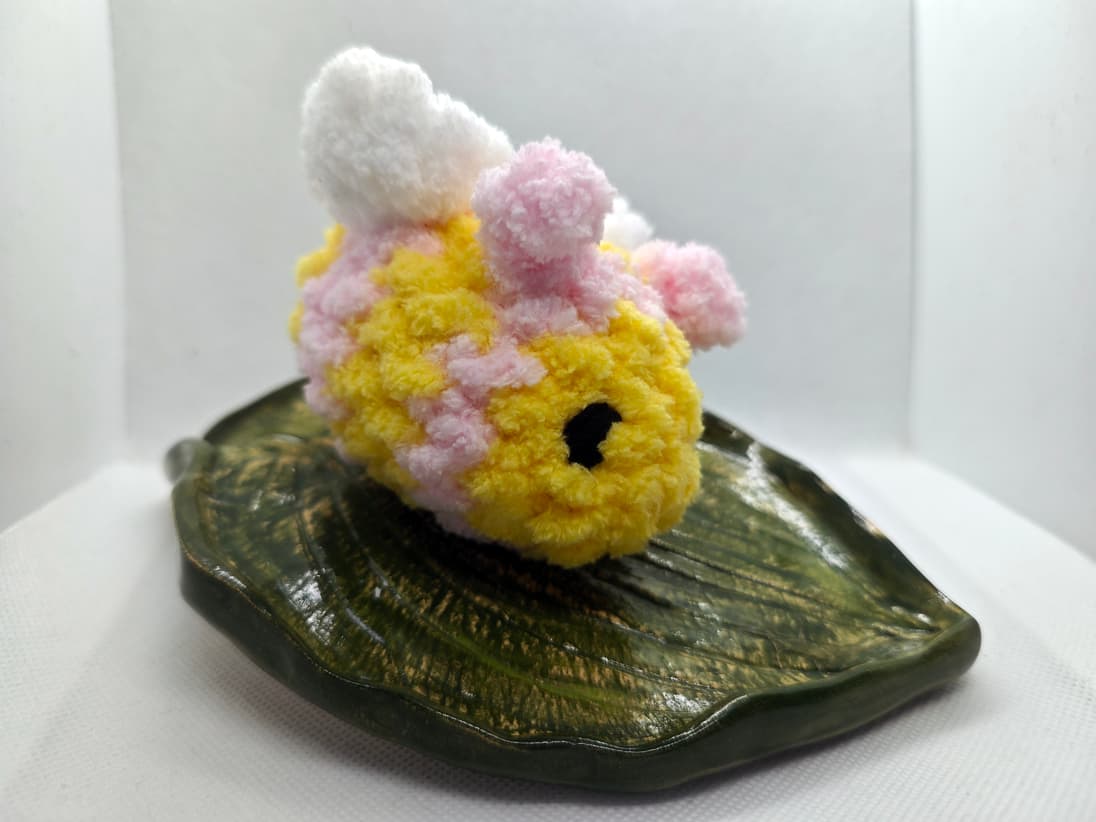 Handmade pastel crochet bee plush toy in yellow and pink with fluffy wings on leaf dish