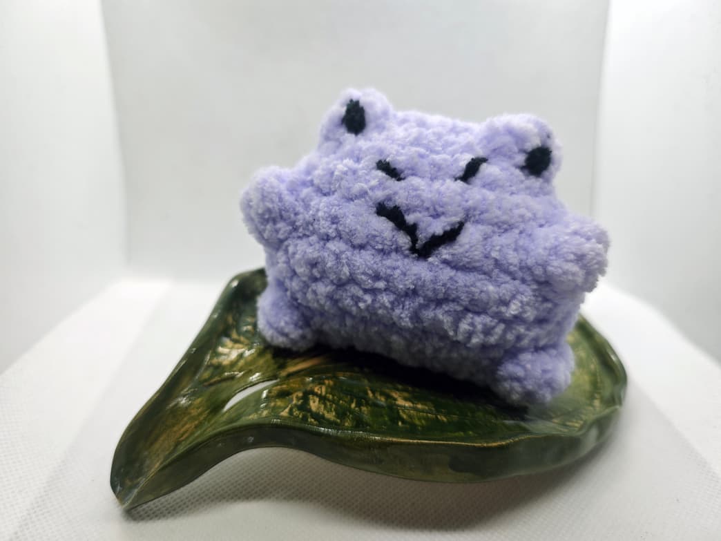 Handmade purple crochet frog plush toy with embroidered face on decorative leaf dish
